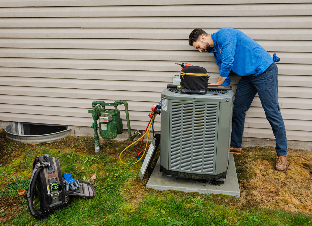 An HVAC technician in a blue shirt servicing an outdoor AC unit
